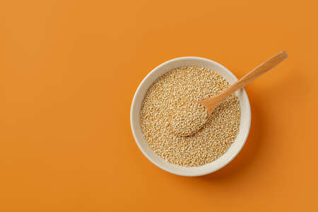 White quinoa seeds and wooden spoon in a beige bowl over orange background. Chenopodium quinoa for...