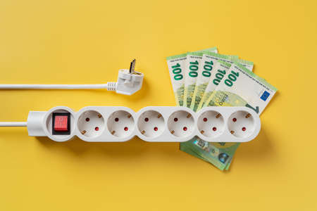 White power strip on 100 euro banknotes and white electric plug over yellow background. Rising energy costs due to power crisis. Increasing lighting, heating and cooling costs while inflation. Top view.の写真素材