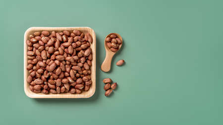 Raw organic peanut on a square wooden plate over green background. Healhy vegetarian snack of whole peeled groundnut seeds. Organic monkey nut as ready to eat dietary nutrition. Copy space. Top view.の写真素材