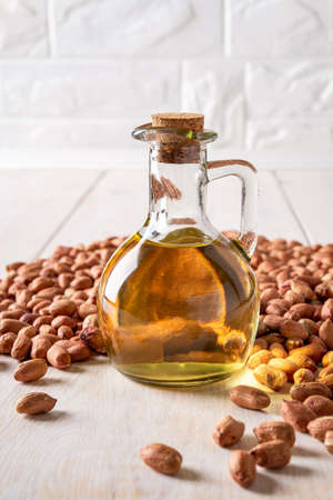 Peanut oil in a glass jug and raw peanuts on a white wooden table. Monounsaturated cooking oil made of Arachis hypogaea. Groundnut as edible seeds and oil crop. Dietary nutrition concept. Front view.の写真素材