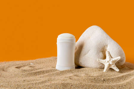 Solid antiperspirant deodorant and sea star near stone on a sand against orange background. Concept...