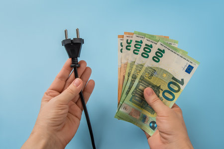 Woman hands hold electric plug and euro banknotes over blue background. High energy costs in Europe. Electric bill increases concept. Soaring of electricity prices. Top view.の写真素材