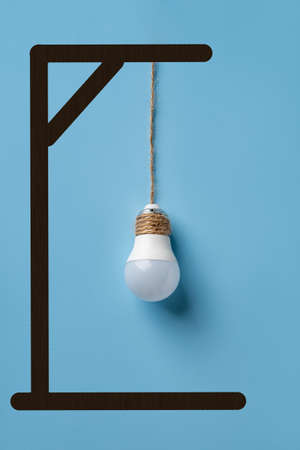 Light bulb hanging from gallows over blue background. Deindustrialization and regression. Reduction of manufacturing, removal of industrial activity concept. Economic crisis. Vertical frame.の写真素材