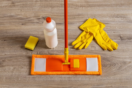 Flat mop and rubber gloves on the floor. Orange microfiber mop, yellow gloves, sponge and liquid cleaner bottle closeup. Mopping floors, homework cleaning routine, housekeeping concept. Top view.の写真素材