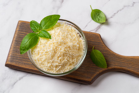Bowl of grated grana padano cheese over marble surface. Finely shredded parmesan in a glass bowl and green basil prepared for cooking. Delicious dairy product. Italian hard cheese. Top view.の写真素材
