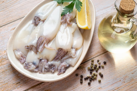 Raw squid fillet on a plate over wooden table. Small calamary tubes, olive oil and lemon prepared...