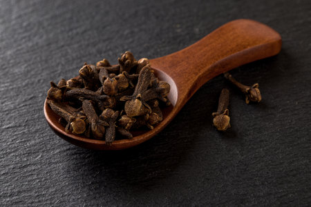 Whole cloves spice in a wooden spoon over black background. Spoon full of dry clove buds close-up. Spice for cooking and herbal medicine concept. Top view.の写真素材
