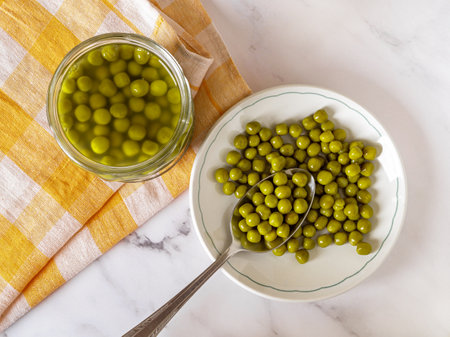 Marinated organic peas on a plate over marble countertop. Canned green peas on a white dish and spoon near glass jar. Vegetable protein, salad ingredient concept. Top view.の写真素材