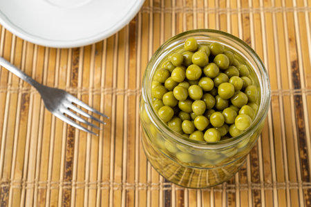 Open jar of canned green peas on a tabletop. Full glass jar of marinated sweet peas, fork and plate. Salad ingredient, vegetable protein, dietary fiber concept. Top view.の写真素材