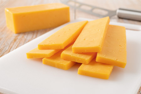 Sliced cheddar cheese close-up. Slaced orange cheese on a white cutting board and knife over rustic...
