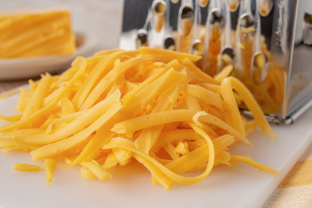 Shredded cheddar cheese close-up. Heap of grated orange cheese and metal grater on a white cutting...