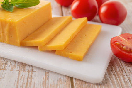 Cheddar cheese slices close-up. Sliced orange cheese on a white cutting board and few red tomatoes...