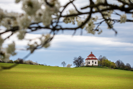 Rural baroque chapel in spring landscape - flowering tree, green field and cloudy sky. Czech Republic, Europe.の写真素材