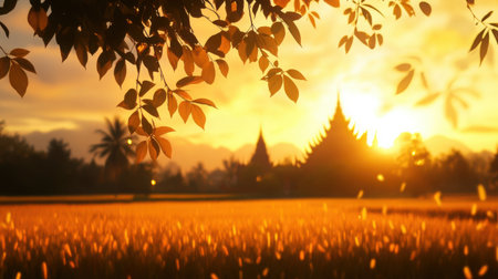 A close-up of leaves rustling in the breeze with a blurred Thai temple in the background and golden rice fields glowing at sunsetの素材