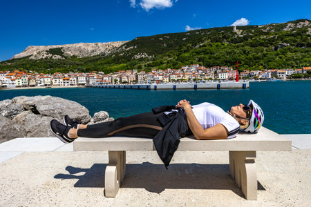 Tired female cyclist rests on a concrete meadow in the center of Baska on the island of KRK, a woman in a bicycle helmet sleeps on a benchの写真素材