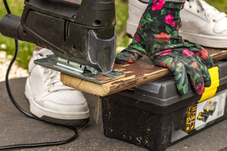 A woman wearing colorful protective gloves cuts boards in the garden using an electric jigsaw....