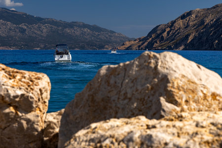 A small tourist boat with an engine sails out of the port in Lopar onto the Adriatic Sea, the boat against the backdrop of a rocky shore and the high Velebit mountains in Dalmatiaの写真素材