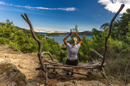 A woman sits on a bench in the middle of a forest, with a panoramic view of Strucic Beach on the island of Rab. Croatiaの写真素材