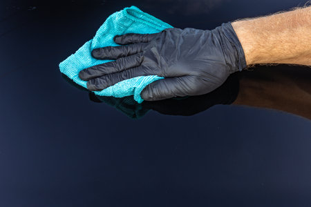 Wiping car wax from the car body with a blue microfiber cloth, a man's hand in a black protective...