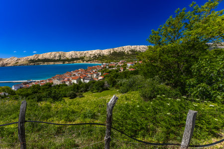 Scenic panoramic view of Baska Bay with turquoise Adriatic Sea, marina, sandy beach, and coastal town surrounded by rocky mountains on Krk Island, Croatia, under a clear blue sky.の写真素材