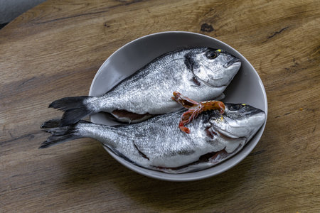 Two fresh raw fish arranged in a ceramic bowl on a wooden table, ready for cooking. The silver...