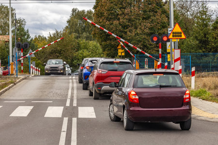 Cars stop at a closed railway crossing while a red and gray diesel locomotive passes through. Safety barriers and warning lights signal the approaching train on a cloudy day.の写真素材