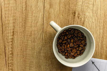 Top view of a coffee cup surrounded by spilled coffee beans on a wooden surface. Symbol of rising coffee prices, inflation, and global market trends in the coffee industry.の写真素材
