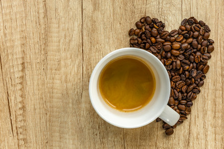 Top view of a cup of coffee with heart-shaped coffee beans on wooden table. Symbol of love for coffee, breakfast, and cozy morning vibes.の写真素材