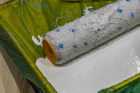 Close up of a paint roller soaked in white paint resting in a green plastic tray. DIY home...
