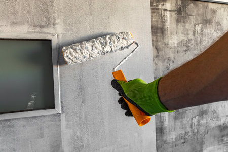 Close up of a hand in a work glove holding a paint roller while painting a beige wall. DIY home renovation, decorating, and interior improvement concept with copy space and natural lightingの写真素材
