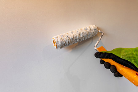 Close up of a hand in a work glove holding a paint roller while painting a beige wall. DIY home renovation, decorating, and interior improvement concept with copy space and natural lightinの写真素材