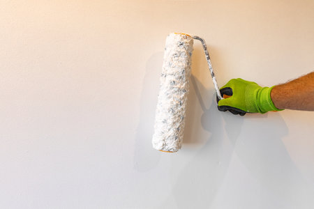 Close up of a hand in a work glove holding a paint roller while painting a beige wall. DIY home renovation, decorating, and interior improvement concept with copy space and natural lightinの写真素材