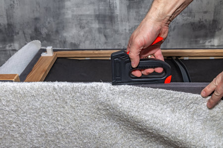 Close up of a black upholstery stapler and metal staples placed on a wooden frame covered with soft white fabric. Concept of furniture renovation, DIY upholstery, and home improvement toolsの写真素材