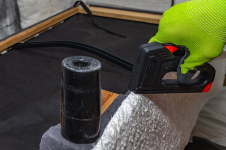 worker's hand in green protective gloves using a black staple gun to attach light fabric to a wooden frame. Upholstery repair or furniture renovation process at home workshopの写真素材