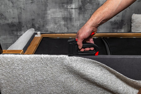 Close up of a black upholstery stapler and metal staples placed on a wooden frame covered with soft white fabric. Concept of furniture renovation, DIY upholstery, and home improvement toolsの写真素材
