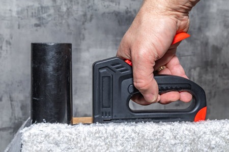 Close up of a black upholstery stapler and metal staples placed on a wooden frame covered with soft...