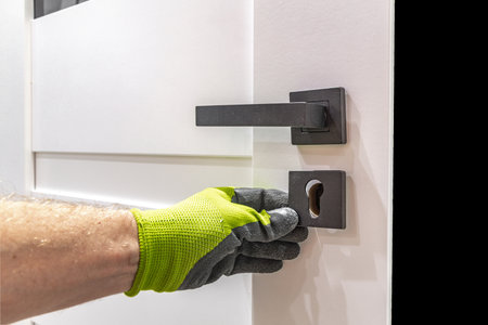 Installing a black door handle with a lock for a white wooden door, framing the door in the frame, working with gloves when installing interior doorsの写真素材