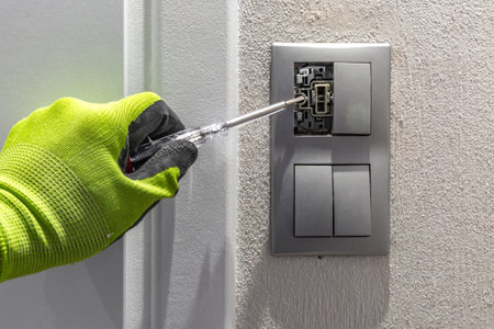 An electrician wearing green protective gloves holds a screwdriver and removes a light switch...