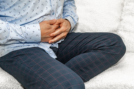 A man in elegant trousers and a long blue shirt sits on a white sofa, holding his stomach, his hands...