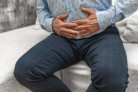 A man in elegant trousers and a long blue shirt sits on a white sofa, holding his stomach, his hands...