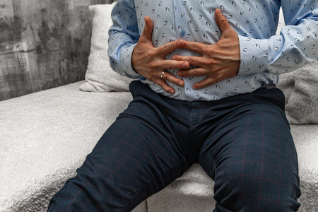 A man in elegant trousers and a long blue shirt sits on a white sofa, holding his stomach, his hands clasped on his stomach.の写真素材