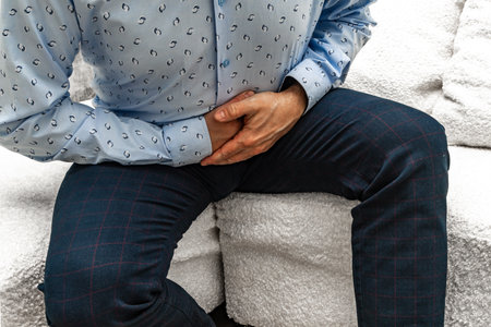 A man in elegant trousers and a long blue shirt sits on a white sofa, holding his stomach, his hands clasped on his stomach.の写真素材