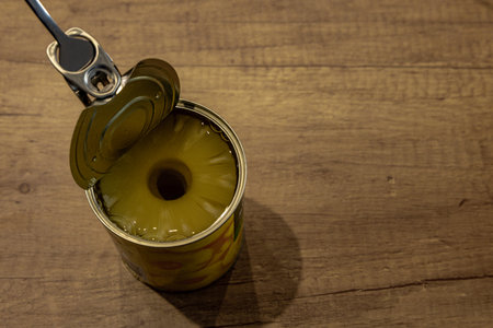Using a fork to remove a pineapple slice from an open can placed on a wooden kitchen tableの写真素材