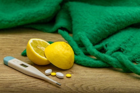 A lemon cut in half, vitamins, painkillers placed on a wooden counter, an electronic thermometer to...