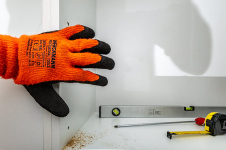 A carpenter wearing protective orange gloves hangs kitchen cabinets, adjusts the hanging of...