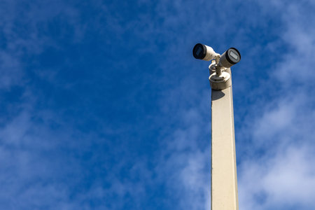 A street surveillance camera mounted on a tall pole against a blue sky, recording in public places,...