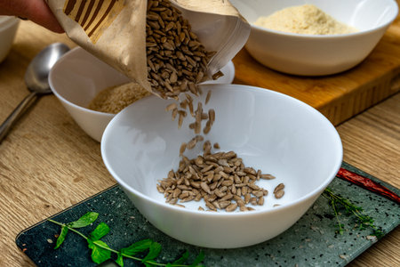 Pouring shelled sunflower seeds into a white porcelain bowl on a wooden kitchen counter, a recipe...