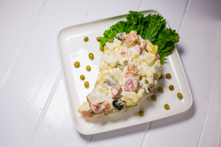 Salad with peas, mayonnaise, potatoes, sausage, cinnamon, carrots and egg in a white plate on a wooden white backgroundの写真素材