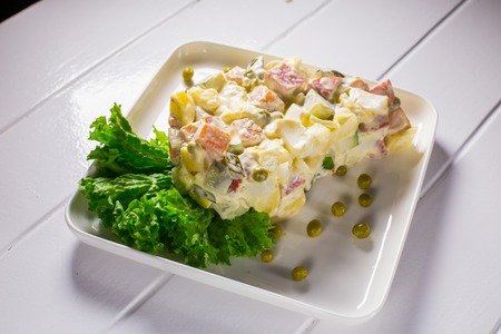 Salad with peas, mayonnaise, potatoes, sausage, cinnamon, carrots and egg in a white plate on a wooden white backgroundの写真素材
