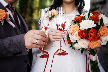 The bride and groom with a wedding bouquet, holding glasses of champagne standing on wedding ceremony under the arch decorated with flowers and greenery of the outdoor.の写真素材